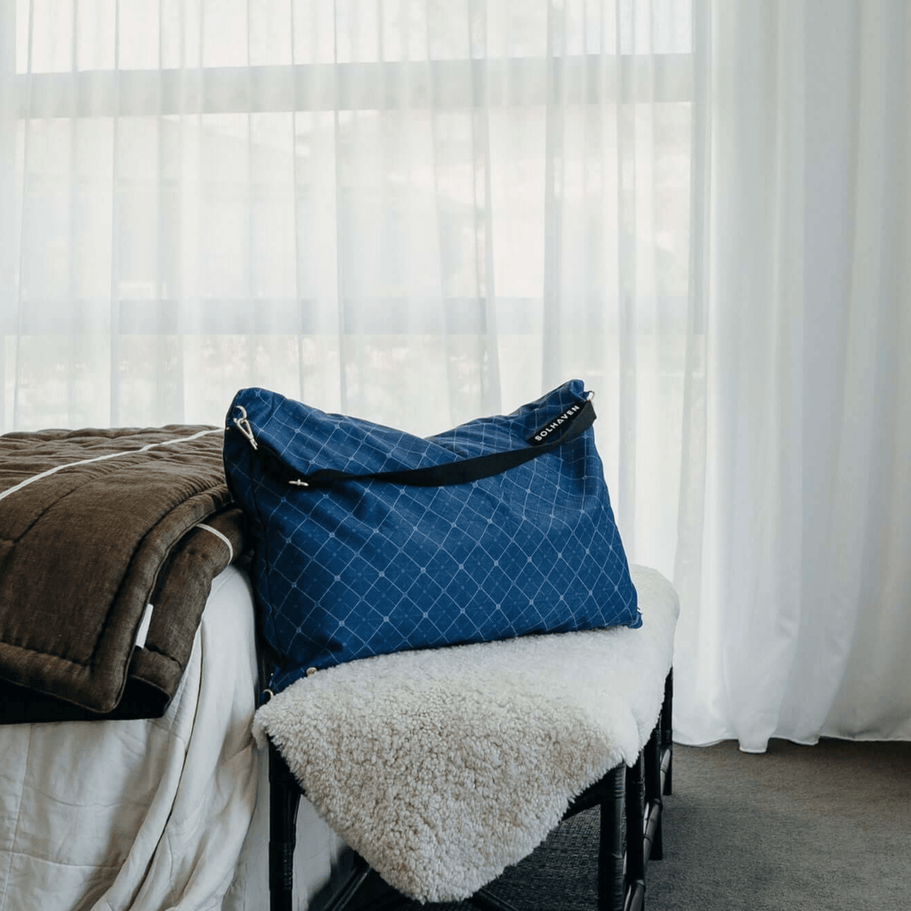 Dream First™ Pillow Bag - Haven - Solhaven sitting on a bench seat in a hotel room with white sheer curtains in the background