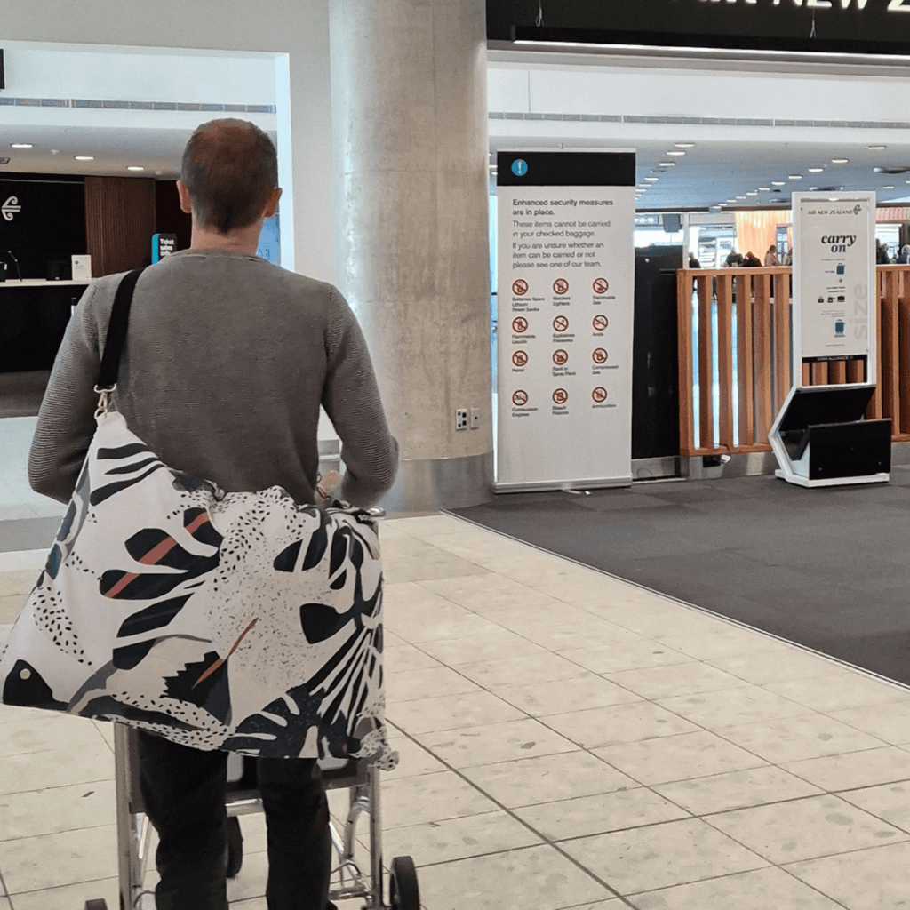 Dream First™ Pillow Bag - Tropic - Solhaven showing a man in a NZ airport with the bag over his shoulder pushing a trolley